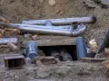 Metalowe rury tworzą skomplikowane przyłącze kanalizacyjne w wykopie. Widać fragmenty izolacji i gruzu.