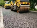 Walec drogowy ugniata świeży asfalt, tworząc gładką nawierzchnię. W tle widać kolejny walec i drzewa.