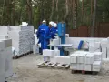 Budowa domu z bloczków betonowych. Pracownicy w niebieskich kombinezonach i białych kaskach przygotowują materiał. Widać stosy bloczków, piłę do cięcia i palety.