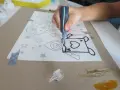 Dziecko tworzy własną **raufazę** na papierze, obrysowując kontury rysunku czarną farbą.