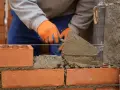 Murarz w pomarańczowych rękawicach układa cegły, używając kielni. To pierwszy krok, jak zrobić grilla z cegły.