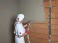 Pracownik w białym stroju nakłada tynki cementowo wapienne na ścianę z pustaków ceramicznych za pomocą agregatu tynkarskiego.