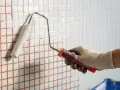 Ręka w rękawicy maluje białą farbą małe, kwadratowe płytki z pomarańczową fugą.