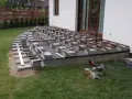 Budowa tarasu kompozytowego: betonowe bloczki podtrzymują legary, tworząc solidną konstrukcję. Widać narzędzia budowlane i materiały.