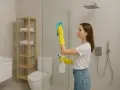 Kobieta w żółtych rękawiczkach czyści szklaną kabinę prysznicową, używając niebieskiej ściereczki i sprayu. Dowiedz się, czym umyć kabinę prysznicową ze szkła, by lśniła czystością.