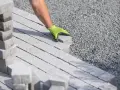 Ręka w zielonej rękawicy układa kostkę brukową na podsypce cementowo piaskowej.