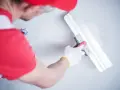 Gipsar w czerwonej czapce i rękawiczkach wygładza ścianę gładzią za pomocą szerokiej pacy.