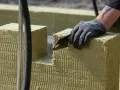 Ręka w rękawicy tnie piłą wełnę skalną 10. Materiał izolacyjny jest cięty na budowie.