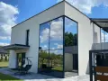 Nowoczesny dom z imponującym, narożnym oknem weneckim, w którym odbija się błękitne niebo z chmurami i zieleń drzew.