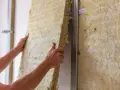 Ręce montują wełnę mineralną do stelaża, pokazując, jak wygłuszyć ścianę. Izolacja akustyczna w budowie.