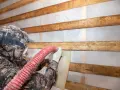 Pracownik w masce i rękawicach aplikuje izolację z **celulozy** za pomocą węża. Drewniane belki tworzą rusztowanie.