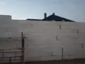 Budowa domu w toku. Ściany z bloczków betonowych czekają na tynkowanie. W tle widać dach i kominy.