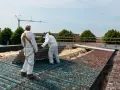 Dwóch pracowników w kombinezonach ochronnych usuwa stare płyty eternitowe z dachu.