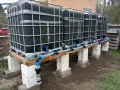 System zbiorników IBC, każdy z czarną osłoną, połączony rurami. Wygląda jak prowizoryczna instalacja do gromadzenia deszczówki, może nawet do nawadniania ogrodu, jak w przypadku systemu mauser.