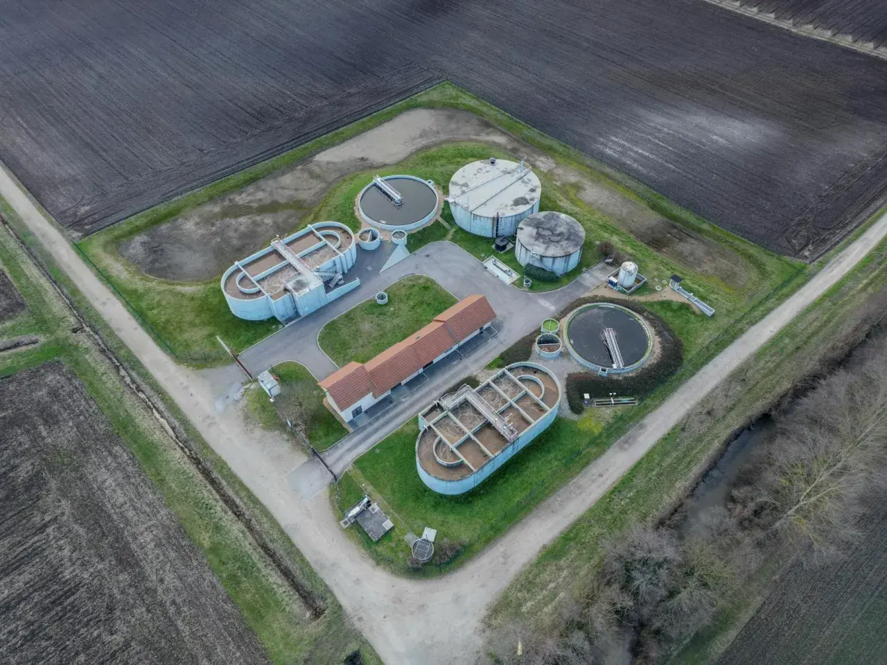 Widok z lotu ptaka na przepompownię ścieków z betonowymi zbiornikami i budynkiem. Otoczona polami uprawnymi.