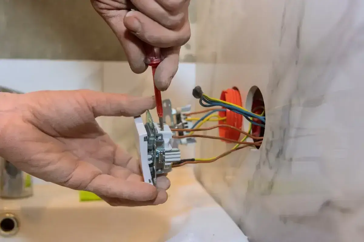 Instalator pokazuje, jak podłączyć gniazdko, przykręcając je do ściany. Widać przewody elektryczne.