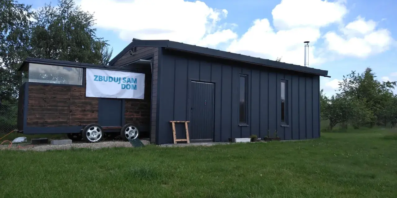 Mobilny garaż bez pozwolenia, z banerem "Zbuduj sam dom", stoi na trawie.