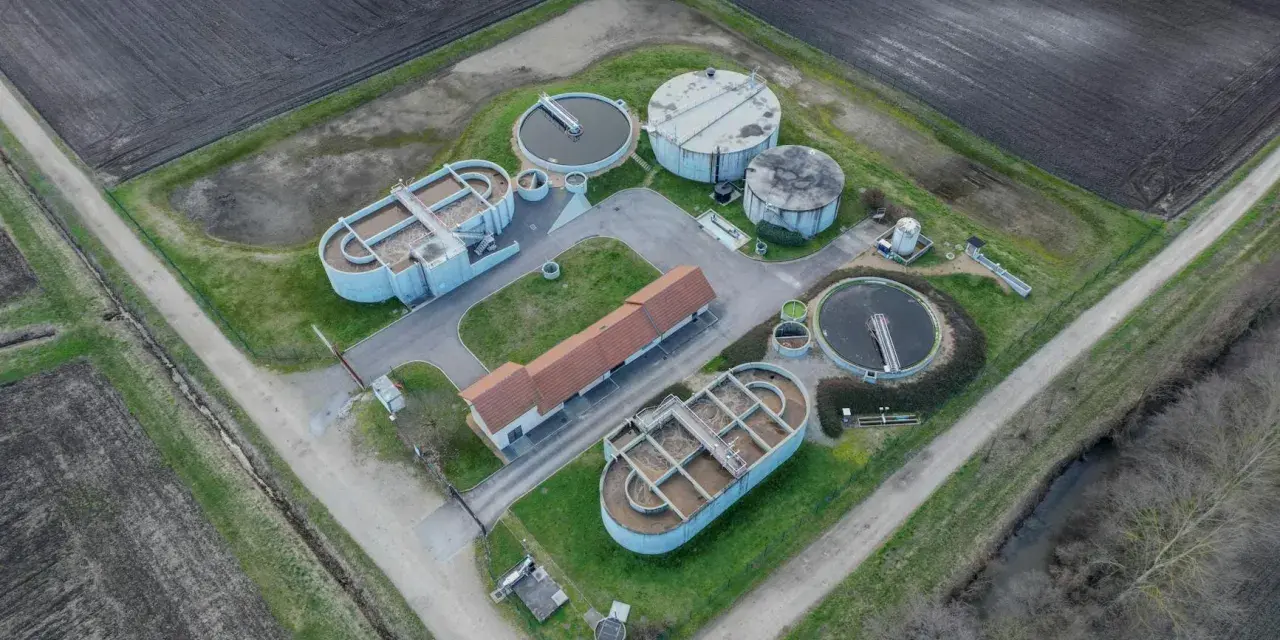 Widok z lotu ptaka na przepompownię ścieków z betonowymi zbiornikami i budynkiem. Otoczona polami uprawnymi.