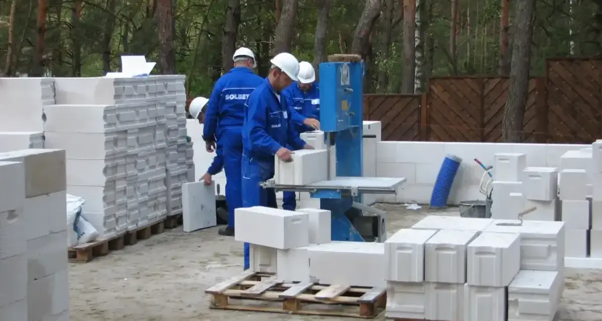 Budowa domu z bloczków betonowych. Pracownicy w niebieskich kombinezonach i białych kaskach przygotowują materiał. Widać stosy bloczków, piłę do cięcia i palety.