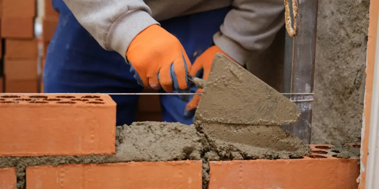 Murarz w pomarańczowych rękawicach układa cegły, używając kielni. To pierwszy krok, jak zrobić grilla z cegły.