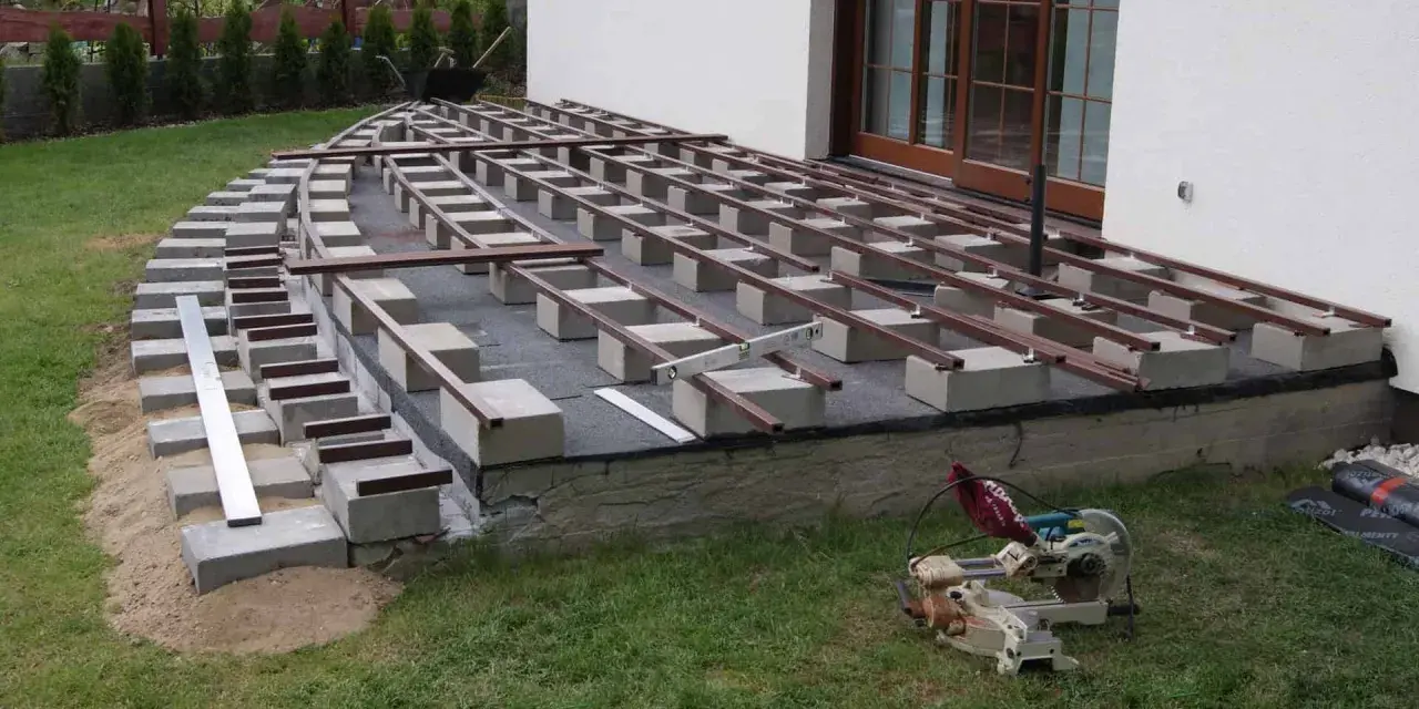 Budowa tarasu kompozytowego: betonowe bloczki podtrzymują legary, tworząc solidną konstrukcję. Widać narzędzia budowlane i materiały.