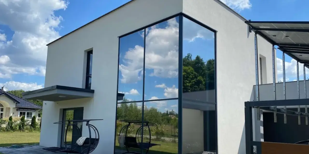 Nowoczesny dom z imponującym, narożnym oknem weneckim, w którym odbija się błękitne niebo z chmurami i zieleń drzew.
