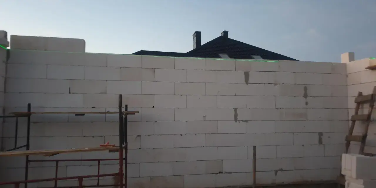 Budowa domu w toku. Ściany z bloczków betonowych czekają na tynkowanie. W tle widać dach i kominy.