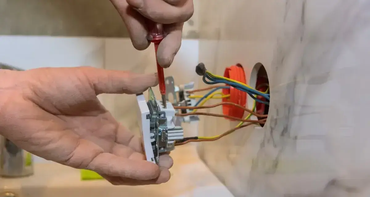 Instalator pokazuje, jak podłączyć gniazdko, przykręcając je do ściany. Widać przewody elektryczne.