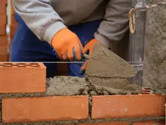 Murarz w pomarańczowych rękawicach układa cegły, używając kielni. To pierwszy krok, jak zrobić grilla z cegły.