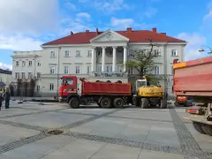 Czerwona wywrotka i żółta koparka na placu przed budynkiem z kolumnami.