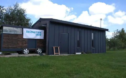 Mobilny garaż bez pozwolenia, z banerem "Zbuduj sam dom", stoi na trawie.