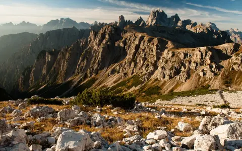 Skaliste szczyty Dolomitów o wschodzie słońca, z kamienistym zboczem i suchą trawą na pierwszym planie.