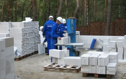 Budowa domu z bloczków betonowych. Pracownicy w niebieskich kombinezonach i białych kaskach przygotowują materiał. Widać stosy bloczków, piłę do cięcia i palety.