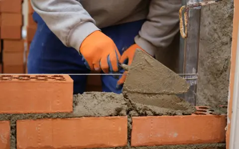 Murarz w pomarańczowych rękawicach układa cegły, używając kielni. To pierwszy krok, jak zrobić grilla z cegły.