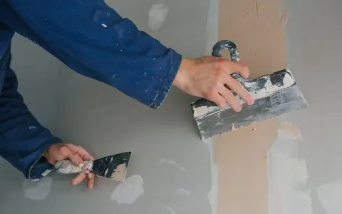 Ręce w niebieskim kombinezonie wykonują szpachlowanie ścian. Jedna ręka trzyma dużą szpachlę, drugą nakłada masę na ścianę.