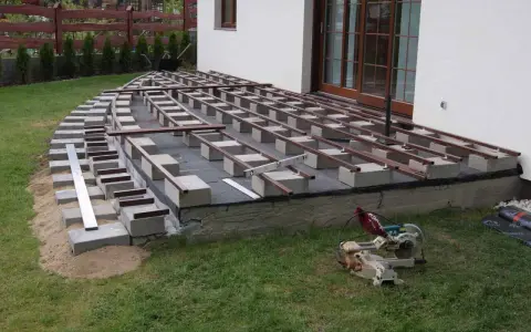 Budowa tarasu kompozytowego: betonowe bloczki podtrzymują legary, tworząc solidną konstrukcję. Widać narzędzia budowlane i materiały.
