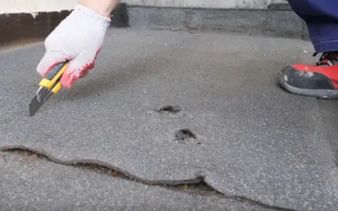 Ręka w rękawicy tnie nożem papę na dachu, przygotowując do naprawy. Widoczne uszkodzenia i pęknięcia.