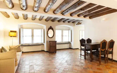 Stary salon z belkami stropowymi i podłogą z terakoty. Kanapa, stół z krzesłami i komoda tworzą przytulną atmosferę.