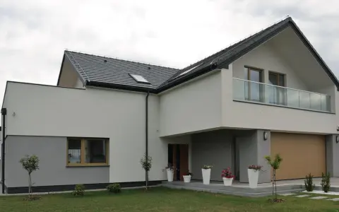 Nowoczesna elewacja budynku z szarymi i białymi tynkami, dużym oknem, balkonem i drewnianą bramą garażową.