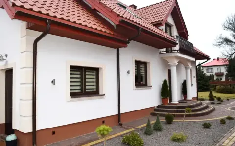Biały dom z brązowymi cokołami, czerwonym dachem i ozdobnymi schodami.