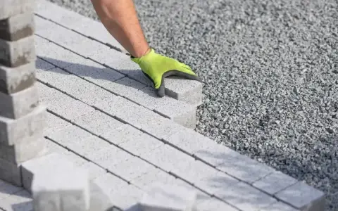 Ręka w zielonej rękawicy układa kostkę brukową na podsypce cementowo piaskowej.