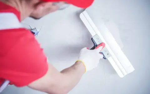 Gipsar w czerwonej czapce i rękawiczkach wygładza ścianę gładzią za pomocą szerokiej pacy.