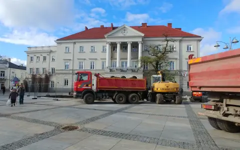 Czerwona wywrotka i żółta koparka na placu przed budynkiem z kolumnami.