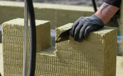 Ręka w rękawicy tnie piłą wełnę skalną 10. Materiał izolacyjny jest cięty na budowie.