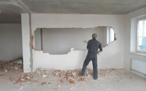 Mężczyzna z młotem burzy ścianę nośną, tworząc otwór. Gruz leży na podłodze.
