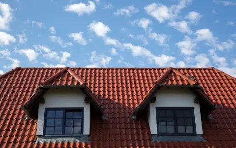 Dachówka ceramiczna w kolorze ceglastym pokrywa dach domu z dwoma lukarnami. Różne rodzaje dachówek tworzą harmonijną całość.