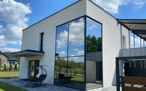 Nowoczesny dom z imponującym, narożnym oknem weneckim, w którym odbija się błękitne niebo z chmurami i zieleń drzew.