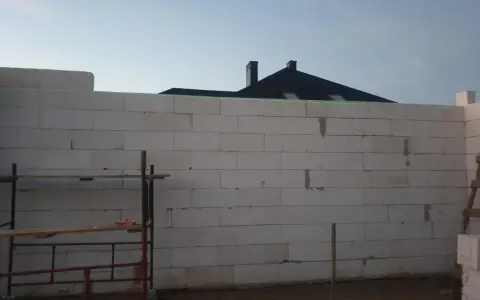 Budowa domu w toku. Ściany z bloczków betonowych czekają na tynkowanie. W tle widać dach i kominy.