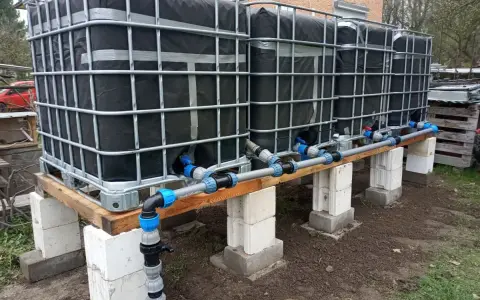 System zbiorników IBC, każdy z czarną osłoną, połączony rurami. Wygląda jak prowizoryczna instalacja do gromadzenia deszczówki, może nawet do nawadniania ogrodu, jak w przypadku systemu mauser.