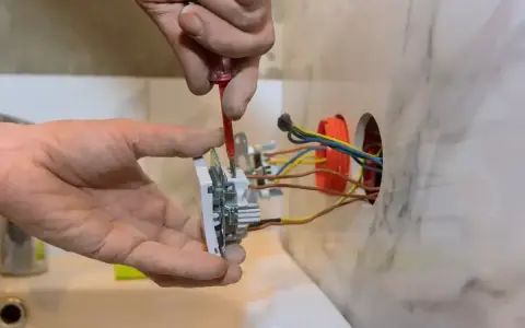 Instalator pokazuje, jak podłączyć gniazdko, przykręcając je do ściany. Widać przewody elektryczne.